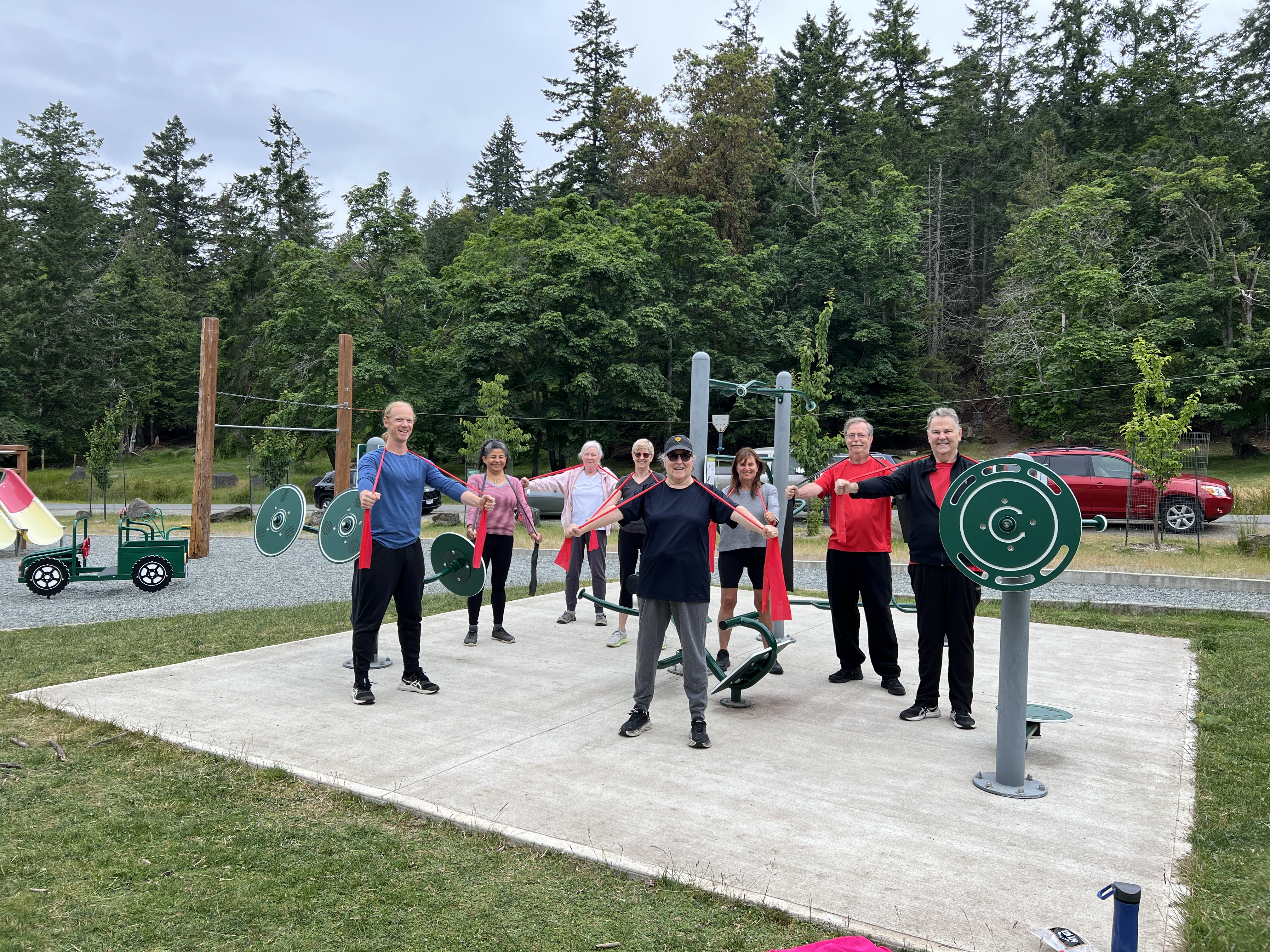 A group of Saturday fitness class participants standing on the outdoor pad at Dinner Bay Park, holding red resistance bands stretched horizontally between their hands.