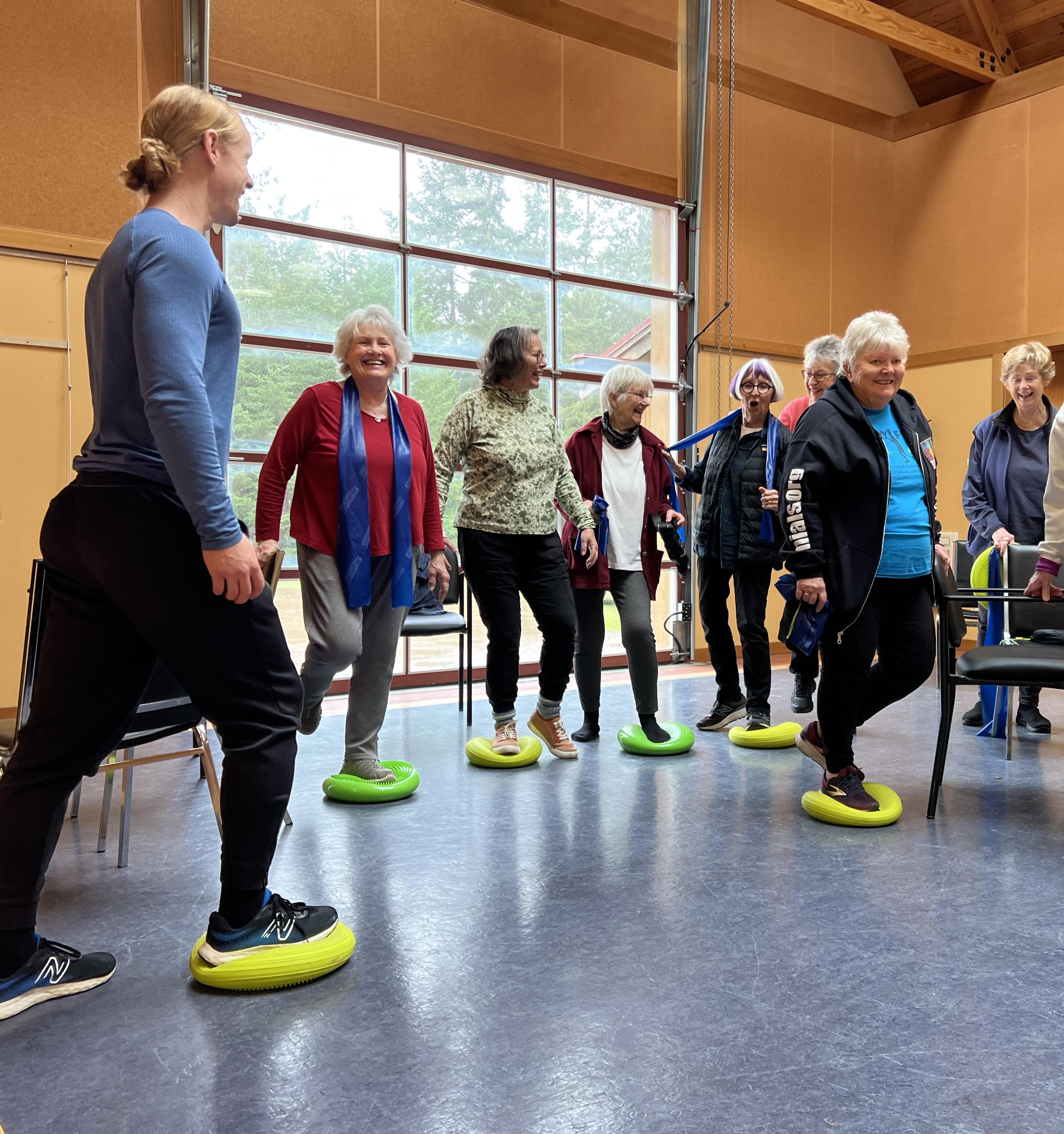 Tim Begley leading a balance class for older adults standing on green balance cushions in a Mayne Island community hall