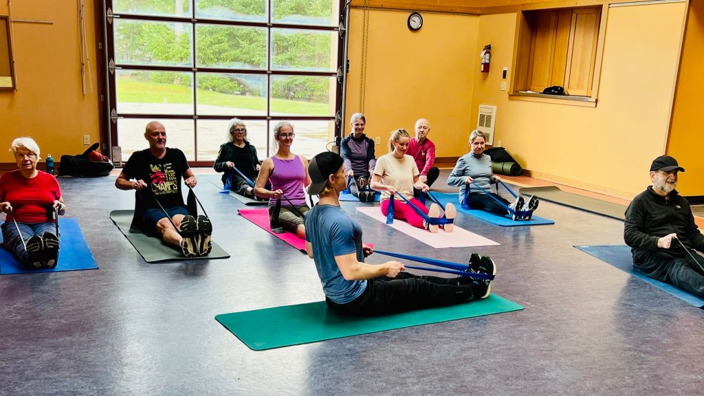 Participants seated on colourful yoga mats inside the Mayne Island Community Centre, doing seated rows with blue resistance bands while the instructor demonstrates in the foreground.